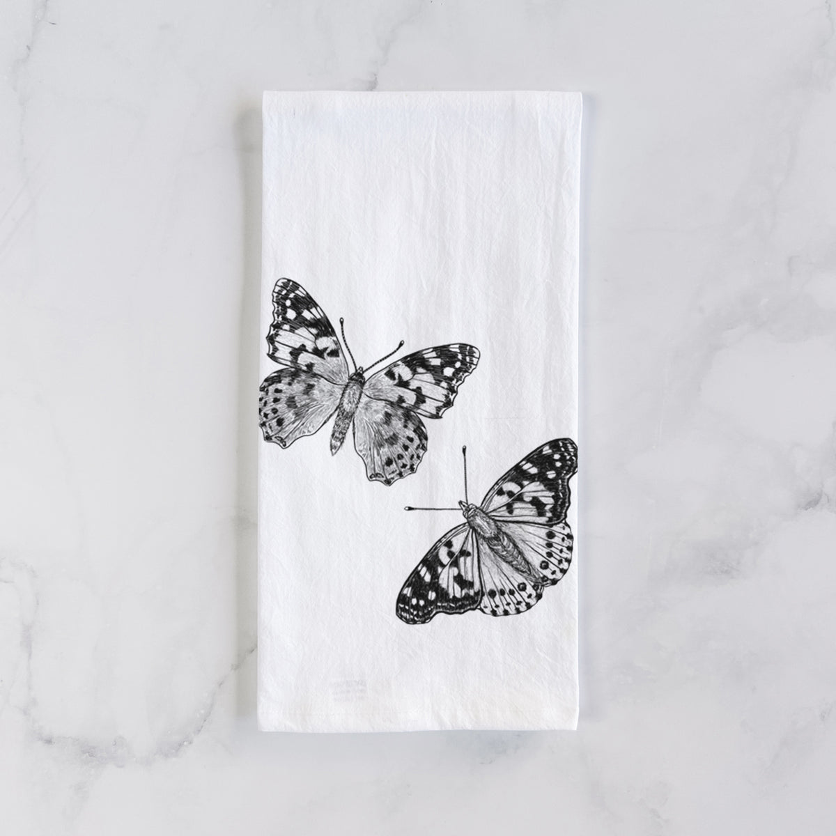 White cotton tea towel laid flat on marble, printed with a detailed black ink illustration of two painted lady butterflies.
