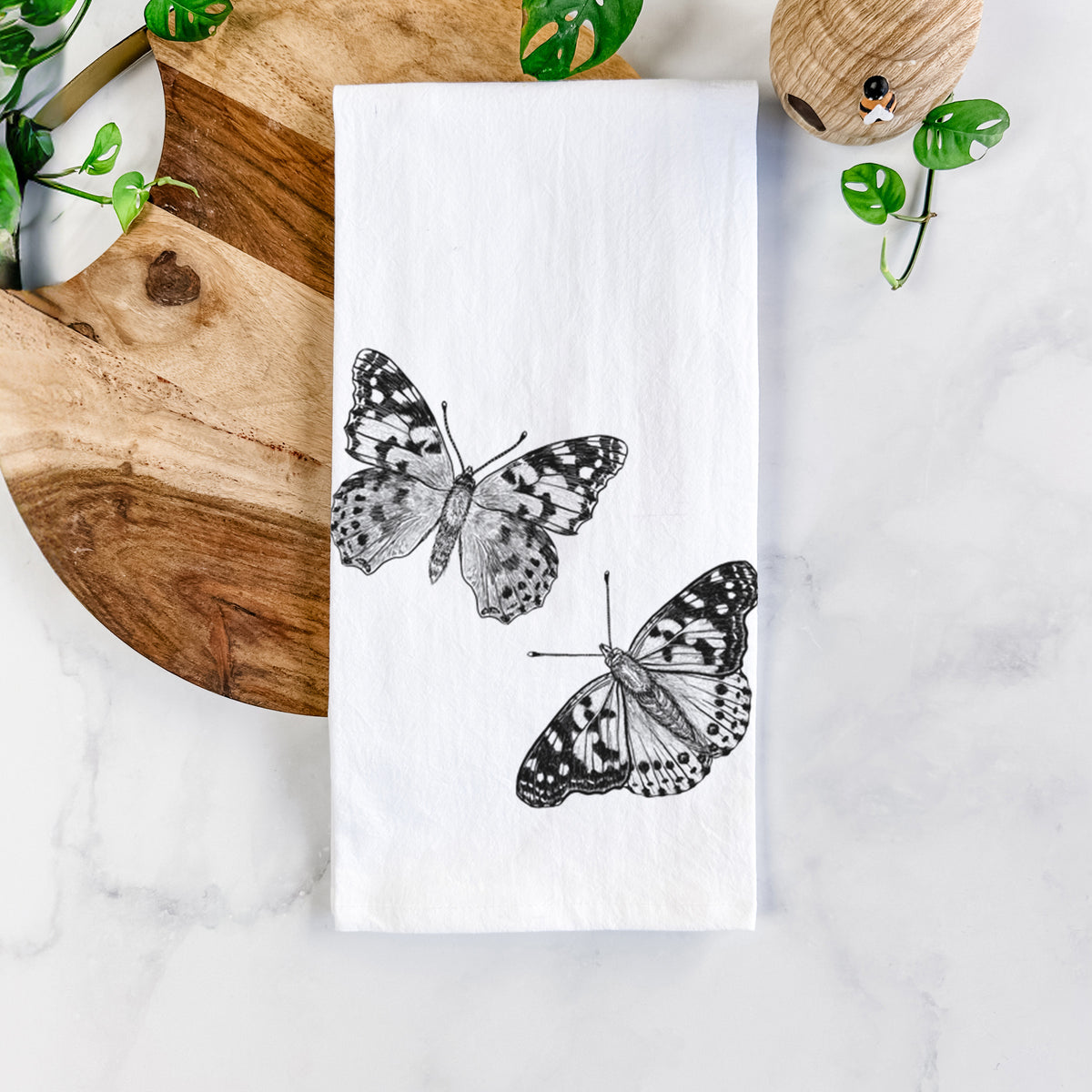 White cotton tea towel printed with a detailed black ink illustration of two painted lady butterflies, on a marble counter beside a round wooden cutting board and greenery.