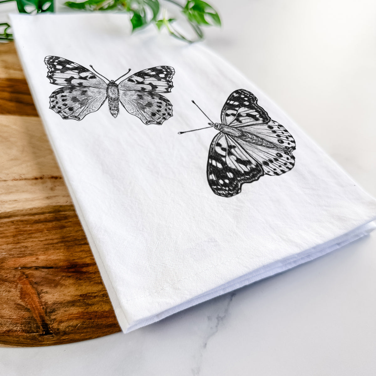Close-up of a folded white tea towel on a wooden board, showing a detailed black ink illustration of two painted lady butterflies.