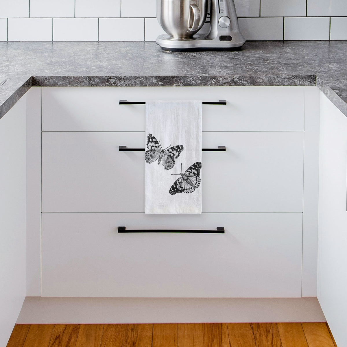 White tea towel printed with a detailed black ink illustration of two painted lady butterflies, hanging from a drawer handle in a bright white kitchen.