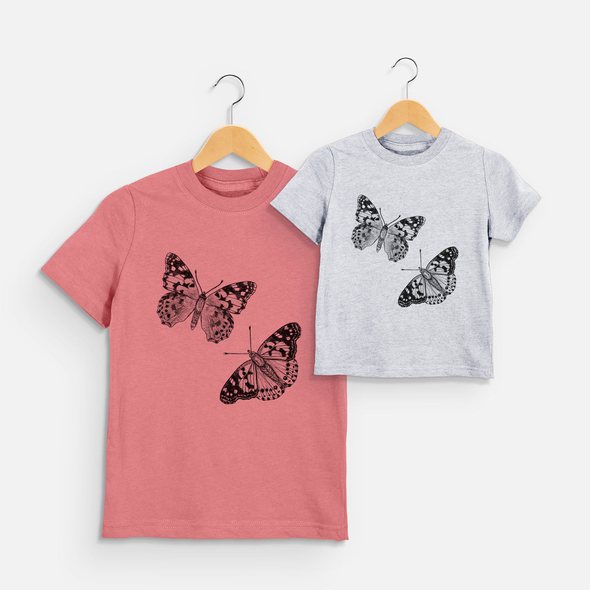 Two youth t-shirts in different colors on hangers, printed with a detailed black ink illustration of two painted lady butterflies.
