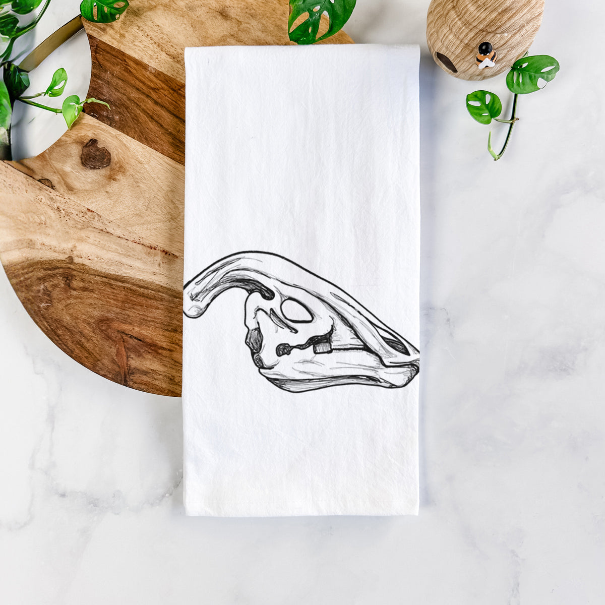 White cotton tea towel printed with a detailed black ink illustration of a crested Parasaurolophus skull, on a marble counter beside a round wooden cutting board and greenery.