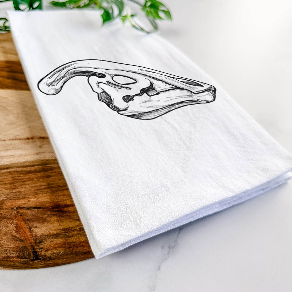 Close-up of a folded white tea towel on a wooden board, showing a detailed black ink illustration of a crested Parasaurolophus skull.