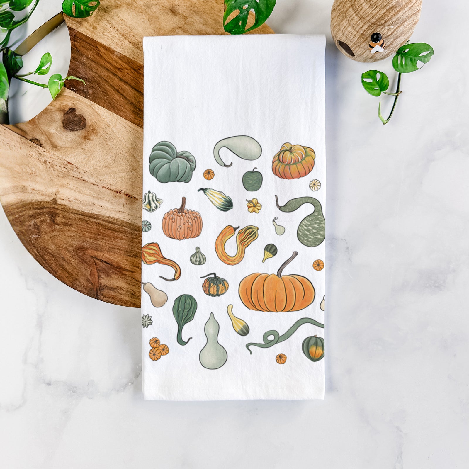 White cotton tea towel printed with a full-color collection of pumpkins, squash and gourds in many shapes and colors, on a marble counter beside a round wooden cutting board and greenery.
