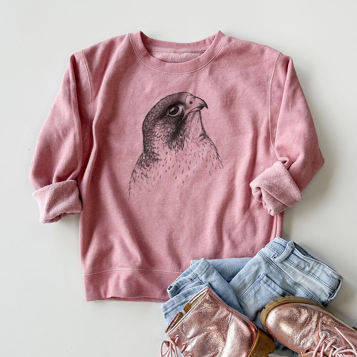 Youth crewneck sweatshirt printed with a detailed black ink portrait of a peregrine falcon's head, styled flat with jeans and boots.