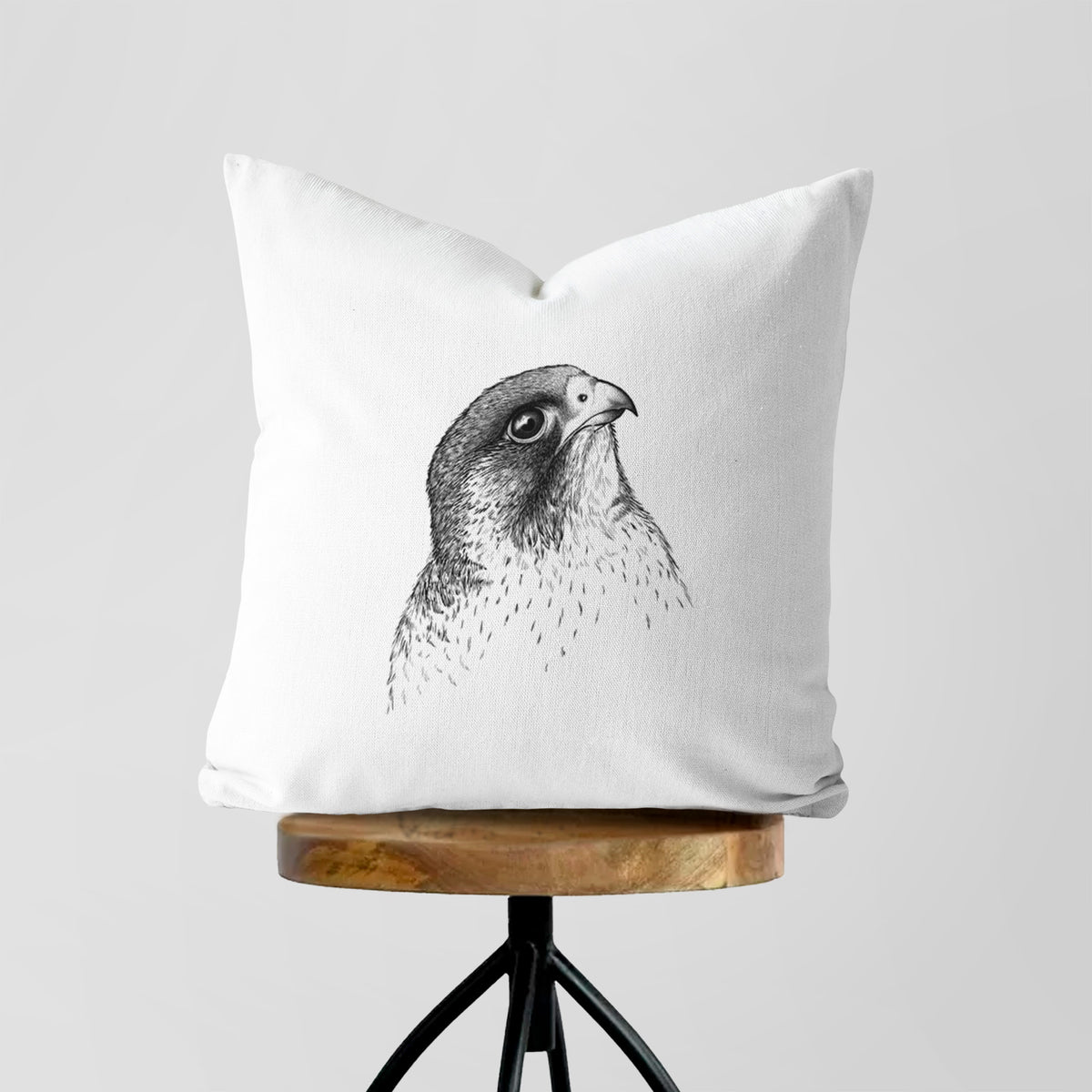 White pillow cover printed with a detailed black ink portrait of a peregrine falcon's head, on a wooden stool.
