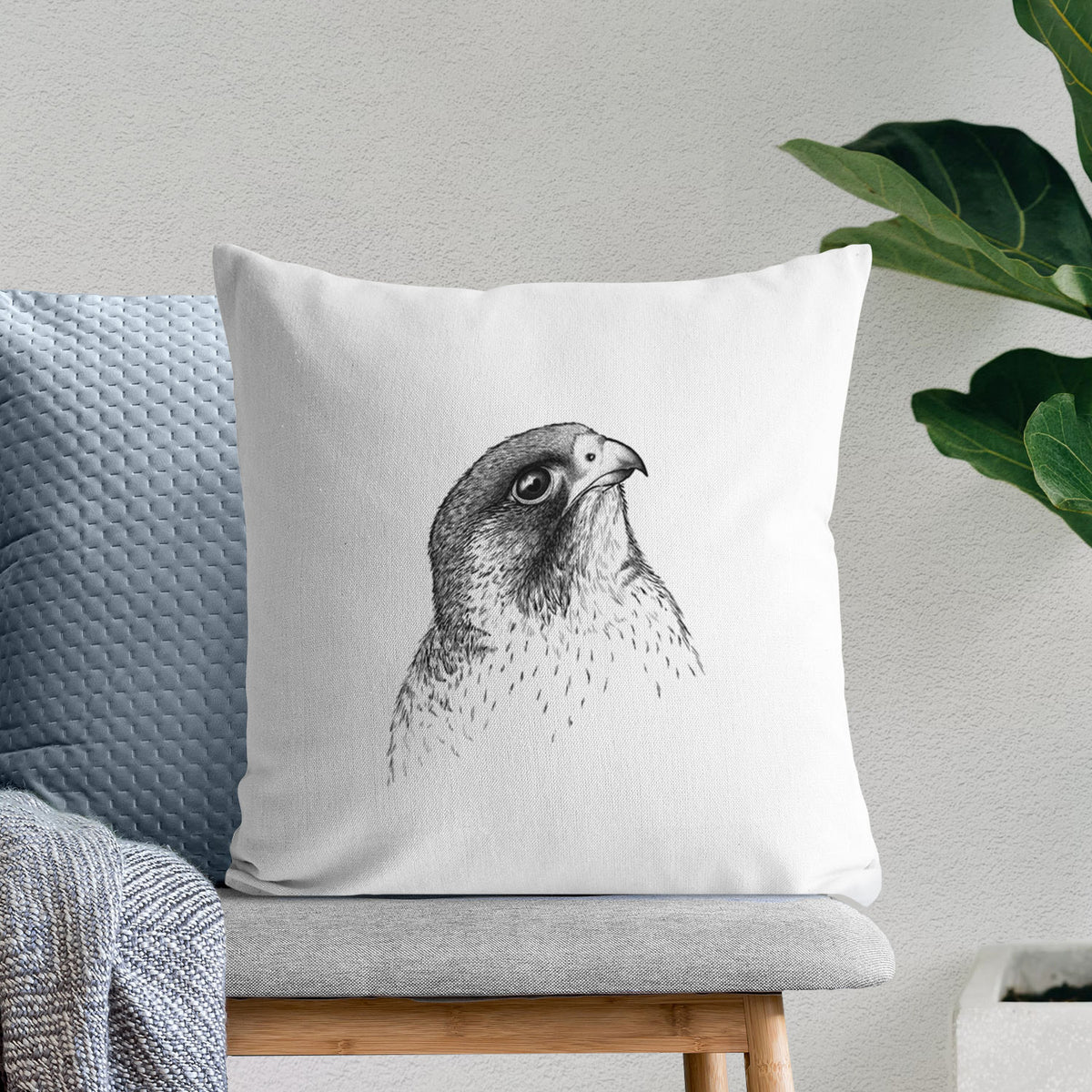 White pillow cover printed with a detailed black ink portrait of a peregrine falcon's head, on a bench beside plants.