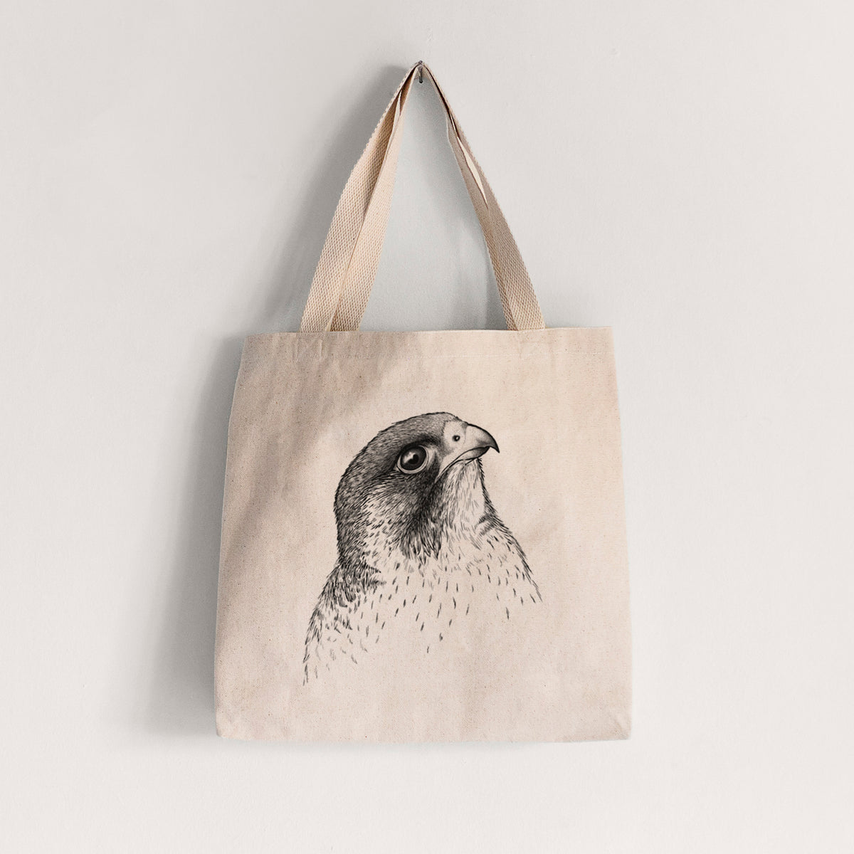 Natural canvas tote bag printed with a detailed black ink portrait of a peregrine falcon's head.
