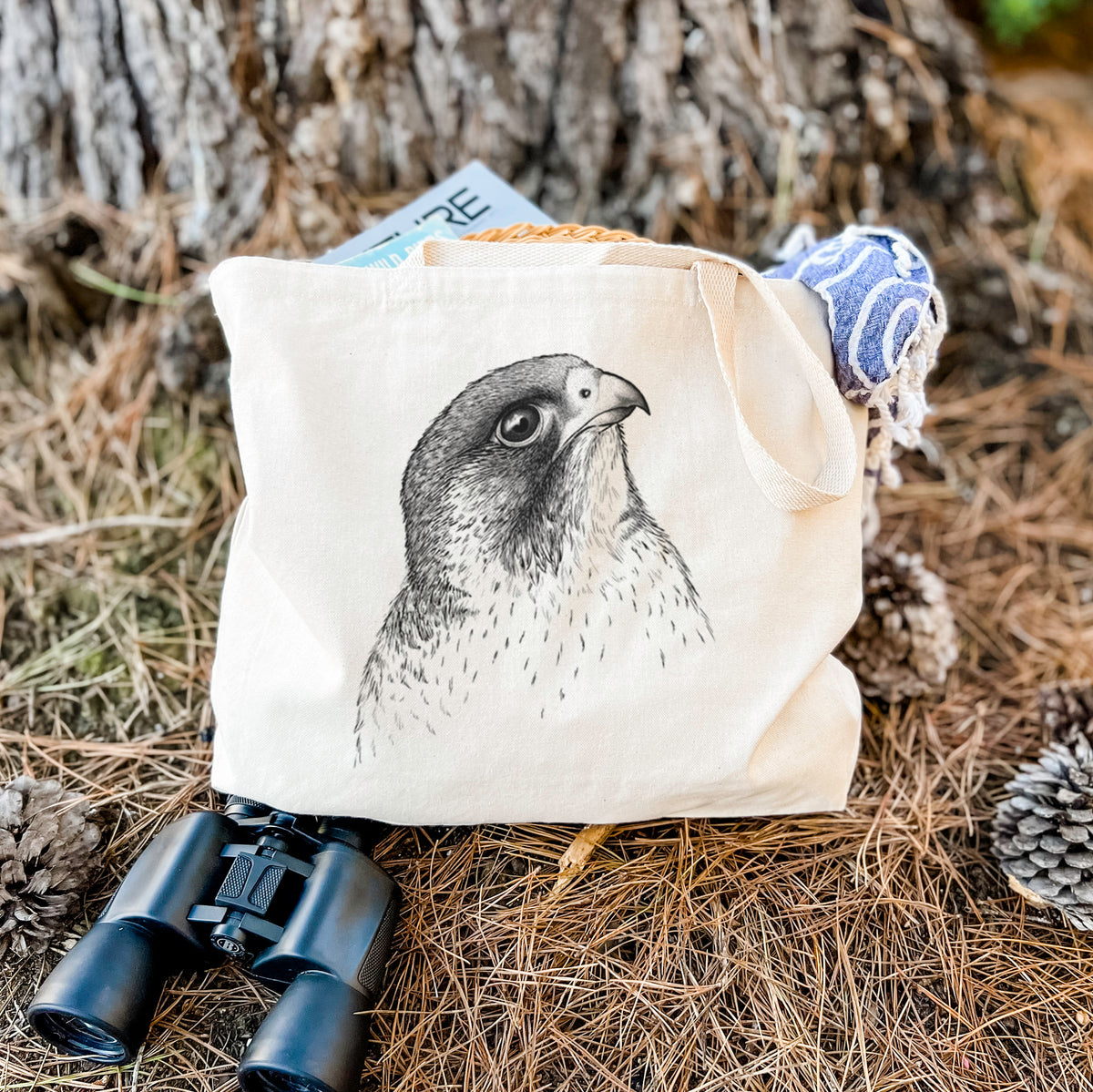 Canvas tote bag printed with a detailed black ink portrait of a peregrine falcon's head, resting on dry grass beside a pair of binoculars.