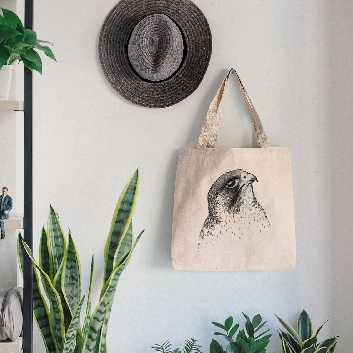 Canvas tote bag printed with a detailed black ink portrait of a peregrine falcon's head, hanging on a wall hook below a hat, next to potted plants.