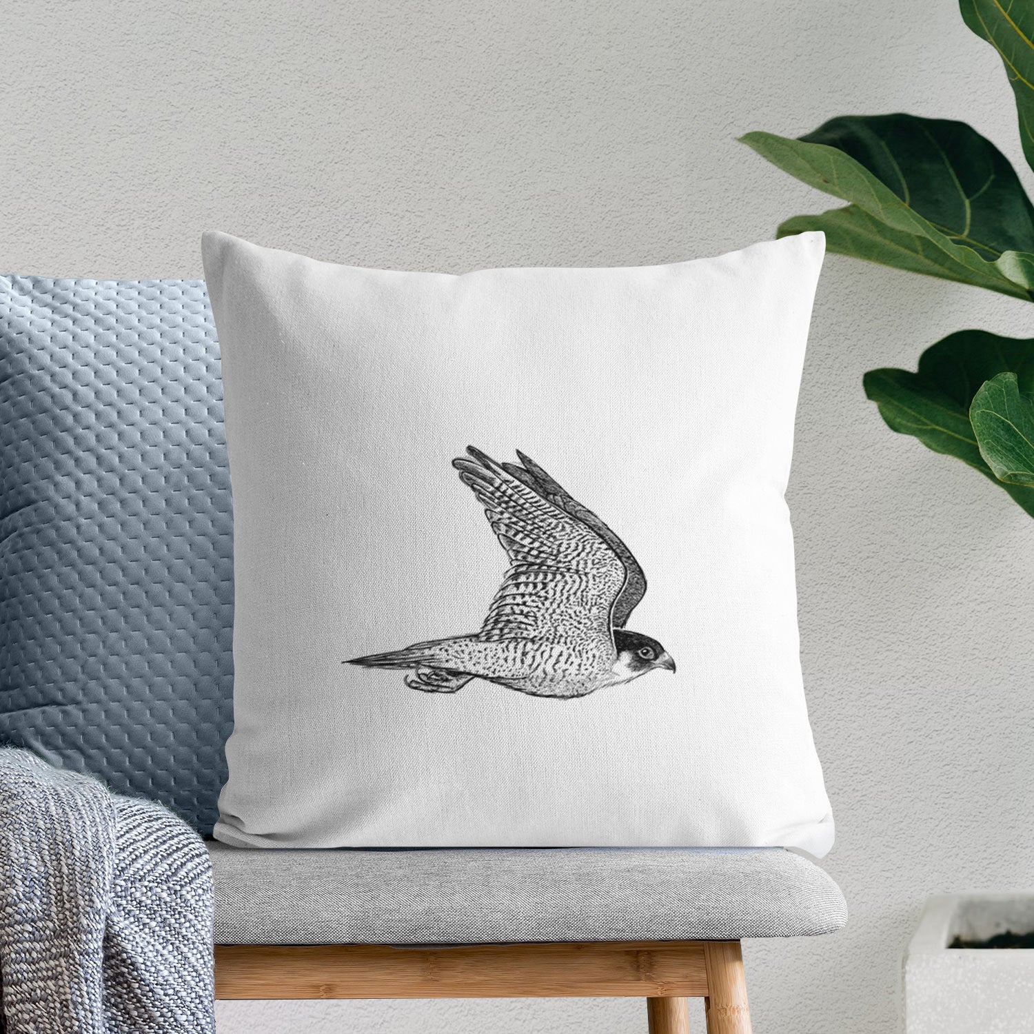 White pillow cover printed with a detailed black ink illustration of a peregrine falcon in flight, on a bench beside plants.