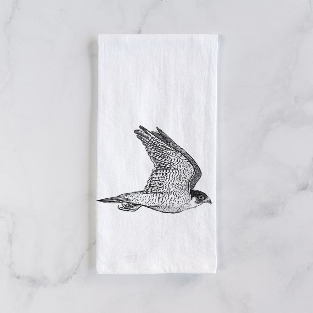 White cotton tea towel laid flat on marble, printed with a detailed black ink illustration of a peregrine falcon in flight.