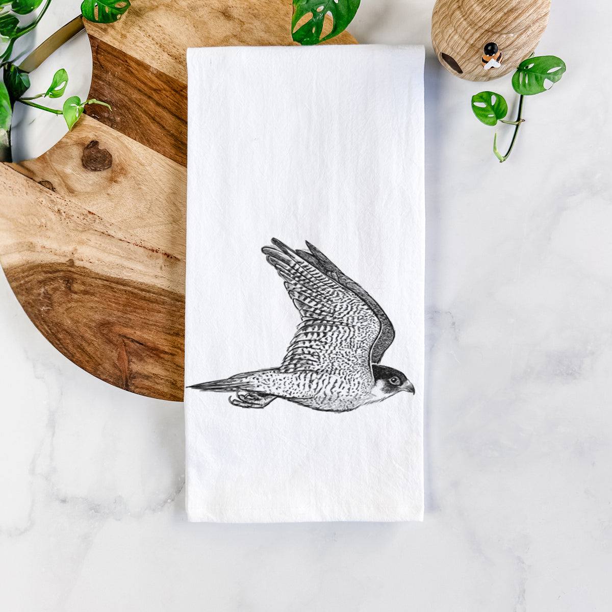 White cotton tea towel printed with a detailed black ink illustration of a peregrine falcon in flight, on a marble counter beside a round wooden cutting board and greenery.