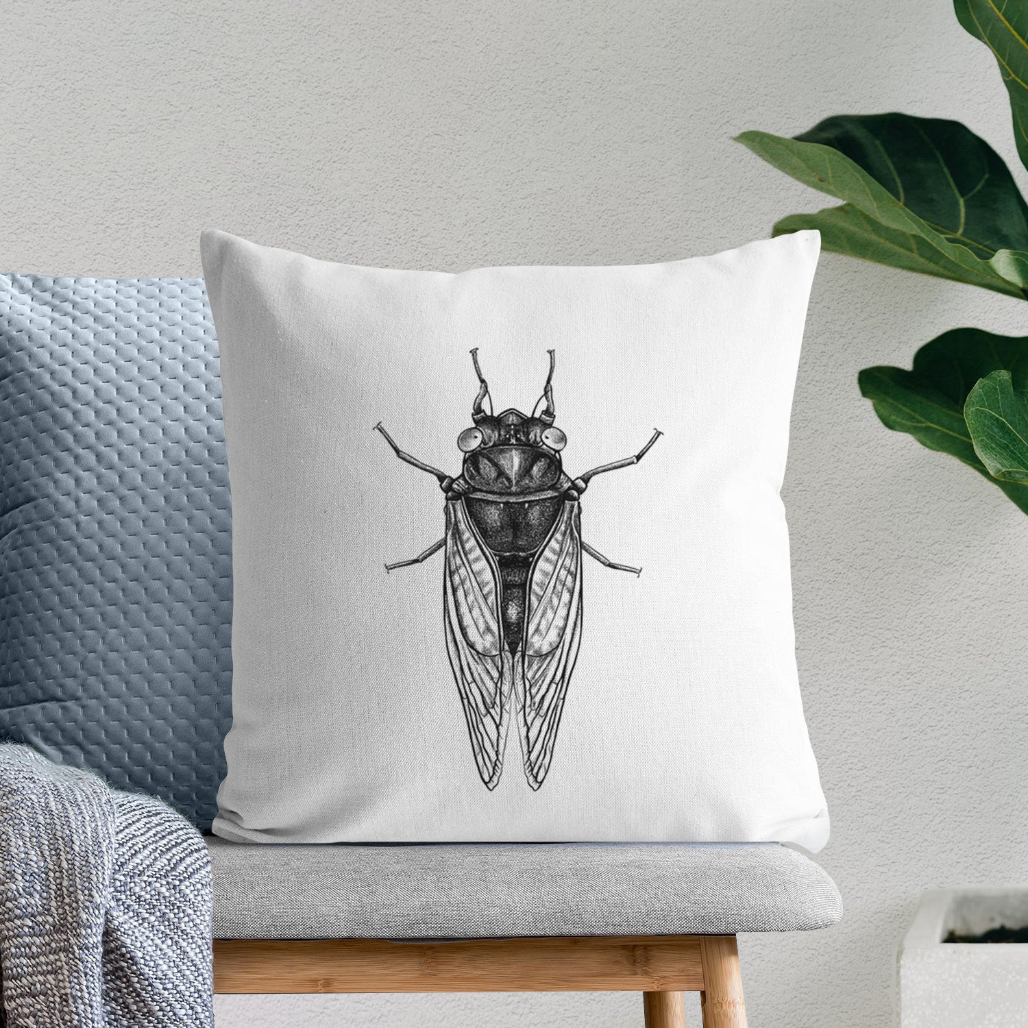White pillow cover printed with a detailed black ink illustration of a periodical cicada with its wings folded, seen from above, on a bench beside plants.