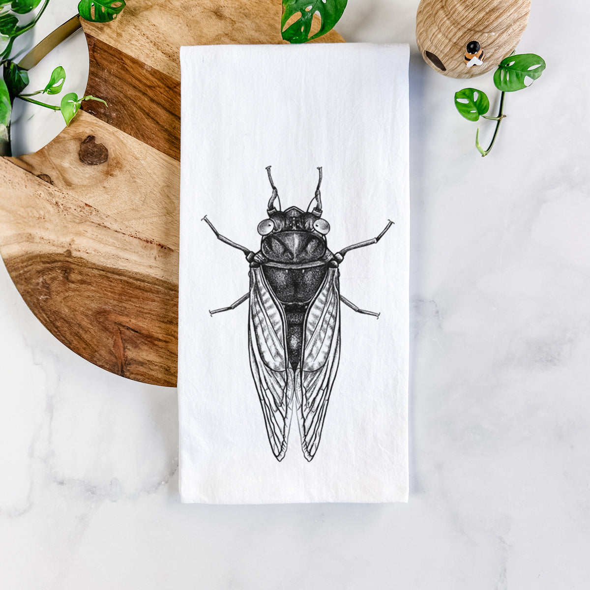 White cotton tea towel printed with a detailed black ink illustration of a periodical cicada with its wings folded, seen from above, on a marble counter beside a round wooden cutting board and greenery.