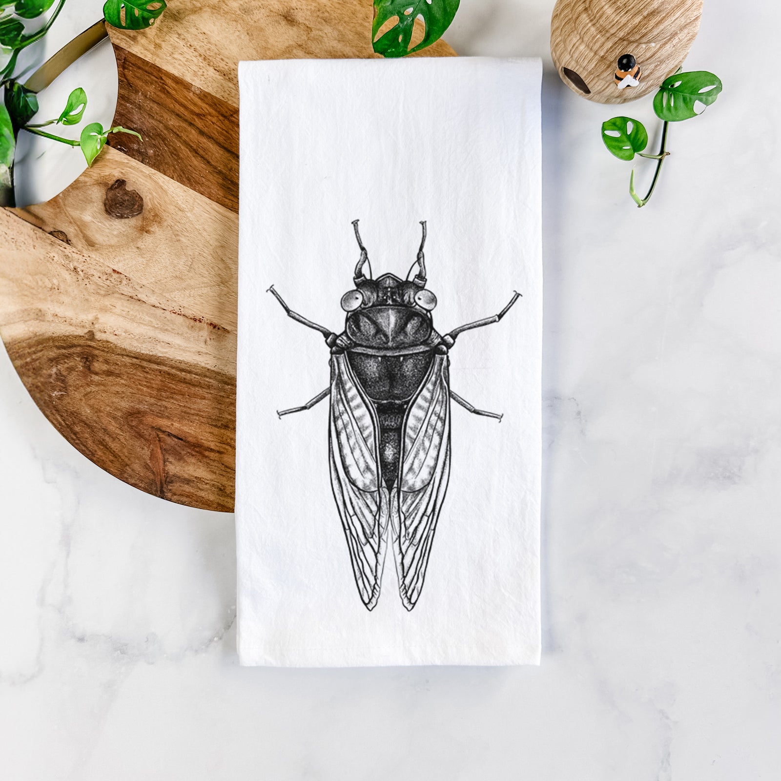 White cotton tea towel printed with a detailed black ink illustration of a periodical cicada with its wings folded, seen from above, on a marble counter beside a round wooden cutting board and greenery.