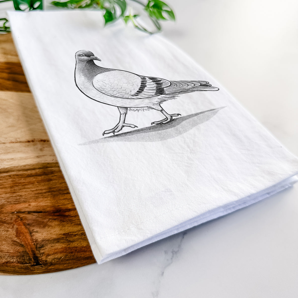 Close-up of a folded white tea towel on a wooden board, showing a black ink illustration of a pigeon standing.