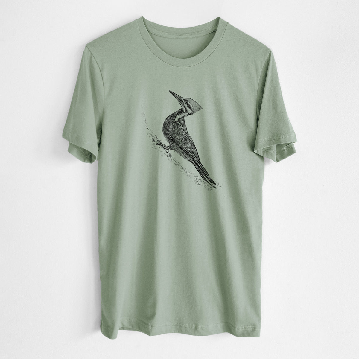 Sage green lightweight cotton t-shirt laid flat, printed with a black ink illustration of a pileated woodpecker clinging to a tree trunk.