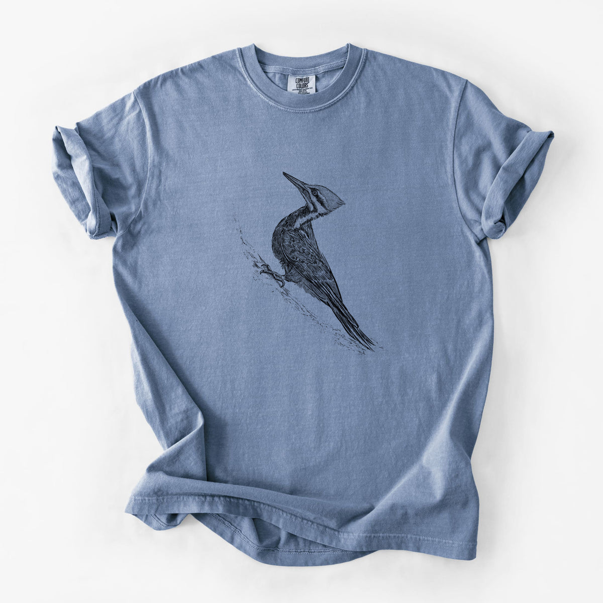 Blue jean Comfort Colors t-shirt laid flat, printed with a black ink illustration of a pileated woodpecker clinging to a tree trunk.