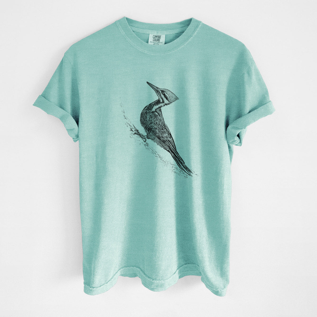 Chalky mint Comfort Colors t-shirt laid flat, printed with a black ink illustration of a pileated woodpecker clinging to a tree trunk.