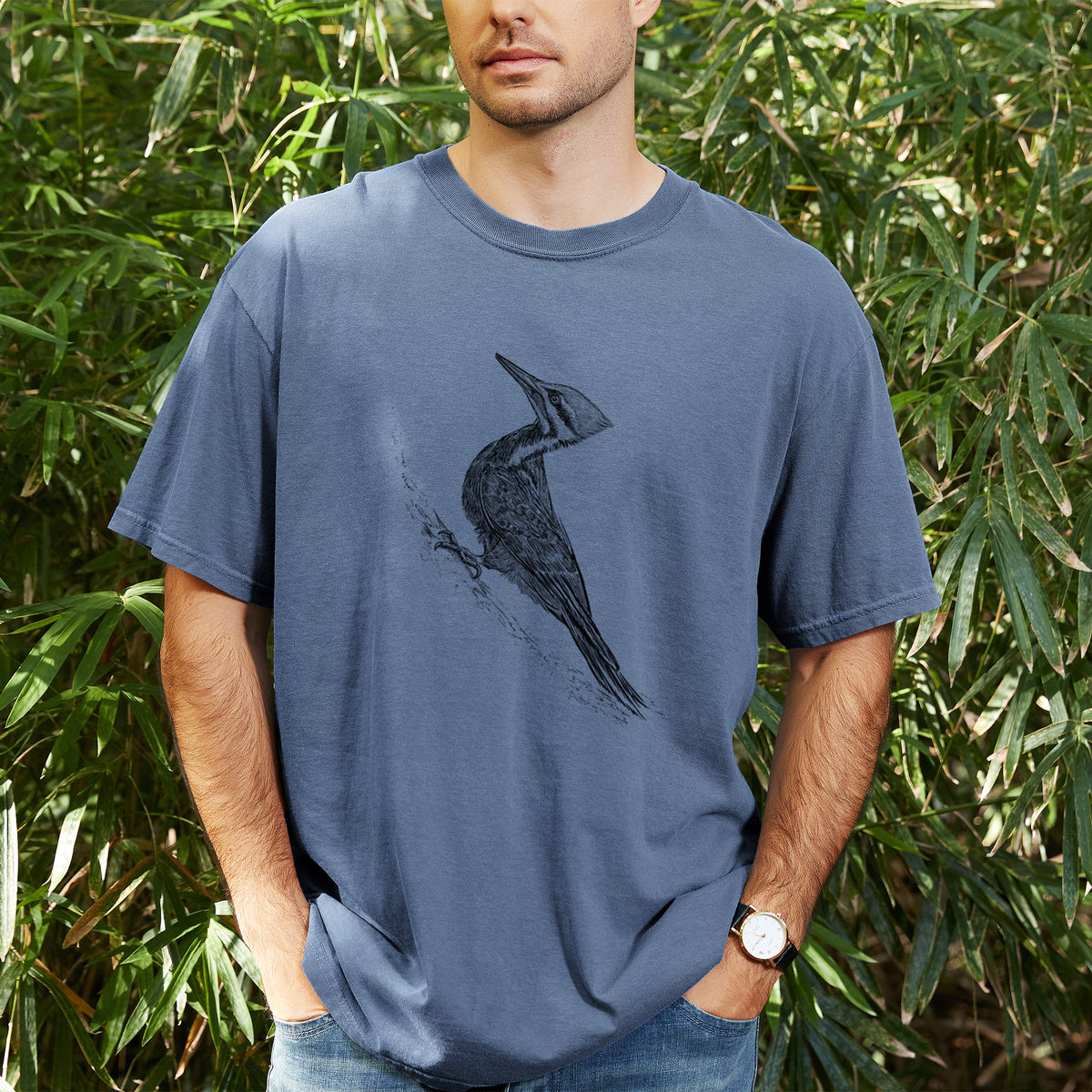 Man wearing a Comfort Colors t-shirt printed with a black ink illustration of a pileated woodpecker clinging to a tree trunk, outdoors.