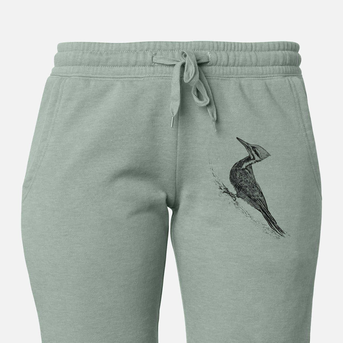 Front of sage green Cali Wave sweatpants, printed with a black ink illustration of a pileated woodpecker clinging to a tree trunk on the leg.