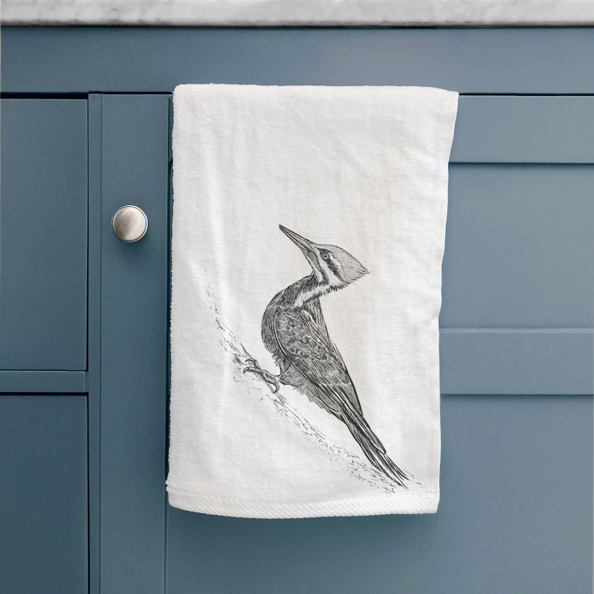 White hand towel printed with a black ink illustration of a pileated woodpecker clinging to a tree trunk, hanging on a cabinet door.