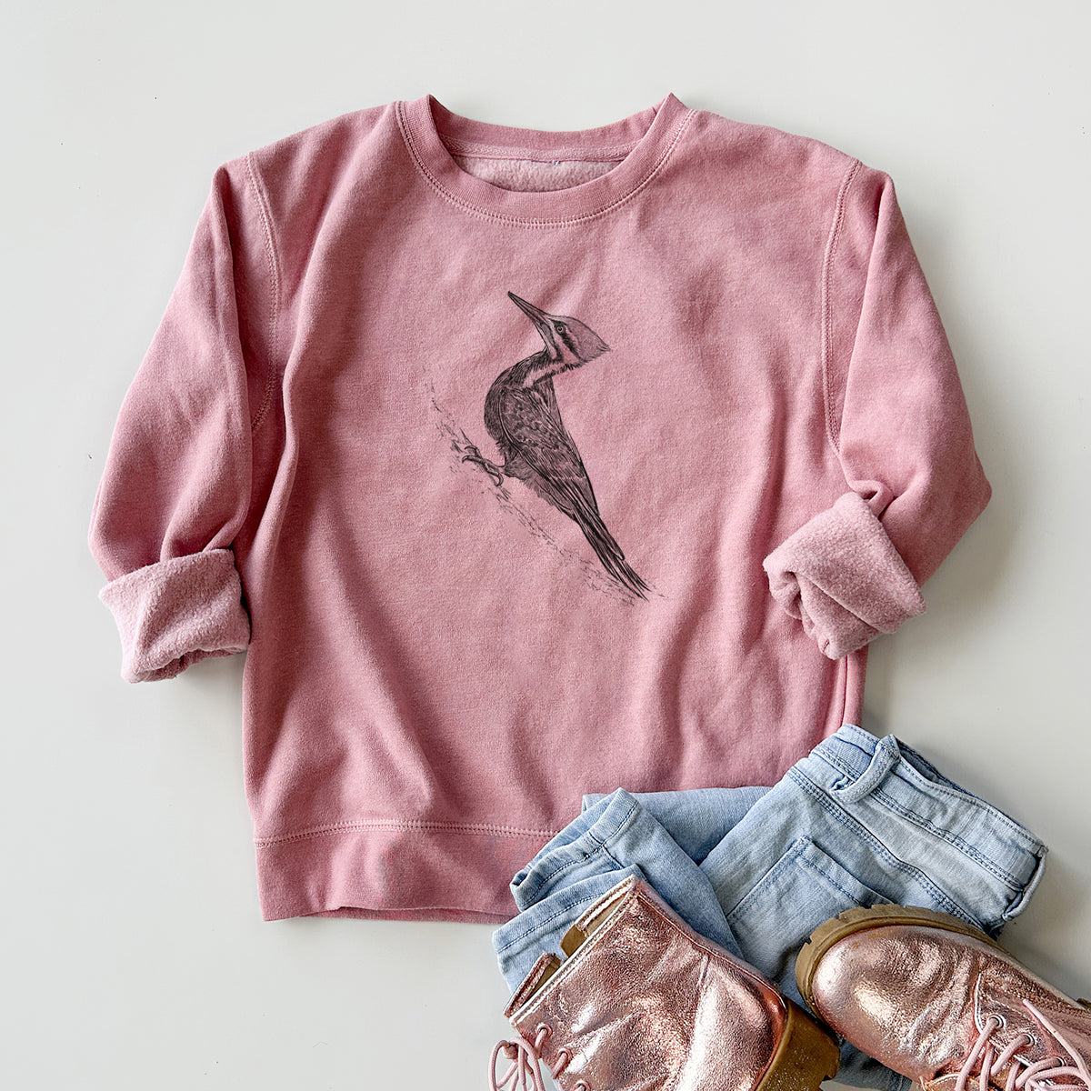 Youth crewneck sweatshirt printed with a black ink illustration of a pileated woodpecker clinging to a tree trunk, styled flat with jeans and boots.