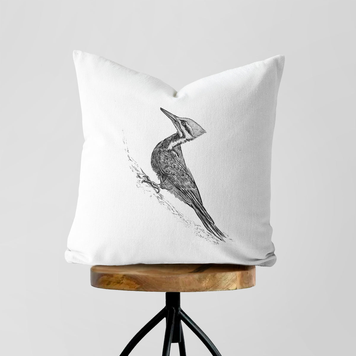 White pillow cover printed with a black ink illustration of a pileated woodpecker clinging to a tree trunk, on a wooden stool.