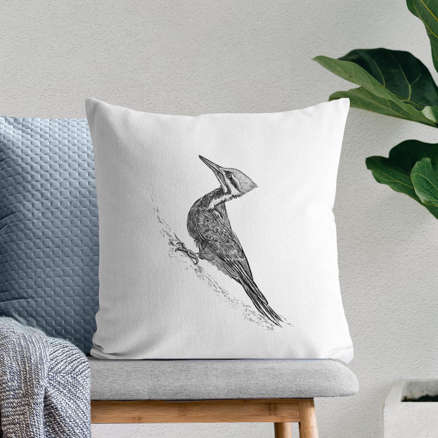 White pillow cover printed with a black ink illustration of a pileated woodpecker clinging to a tree trunk, on a bench beside plants.