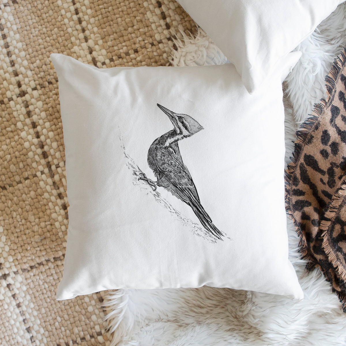 White pillow cover printed with a black ink illustration of a pileated woodpecker clinging to a tree trunk, on a woven chair.