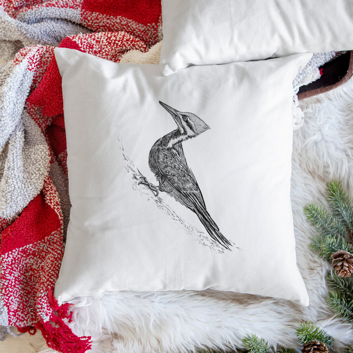 White pillow cover printed with a black ink illustration of a pileated woodpecker clinging to a tree trunk, on a woven chair.