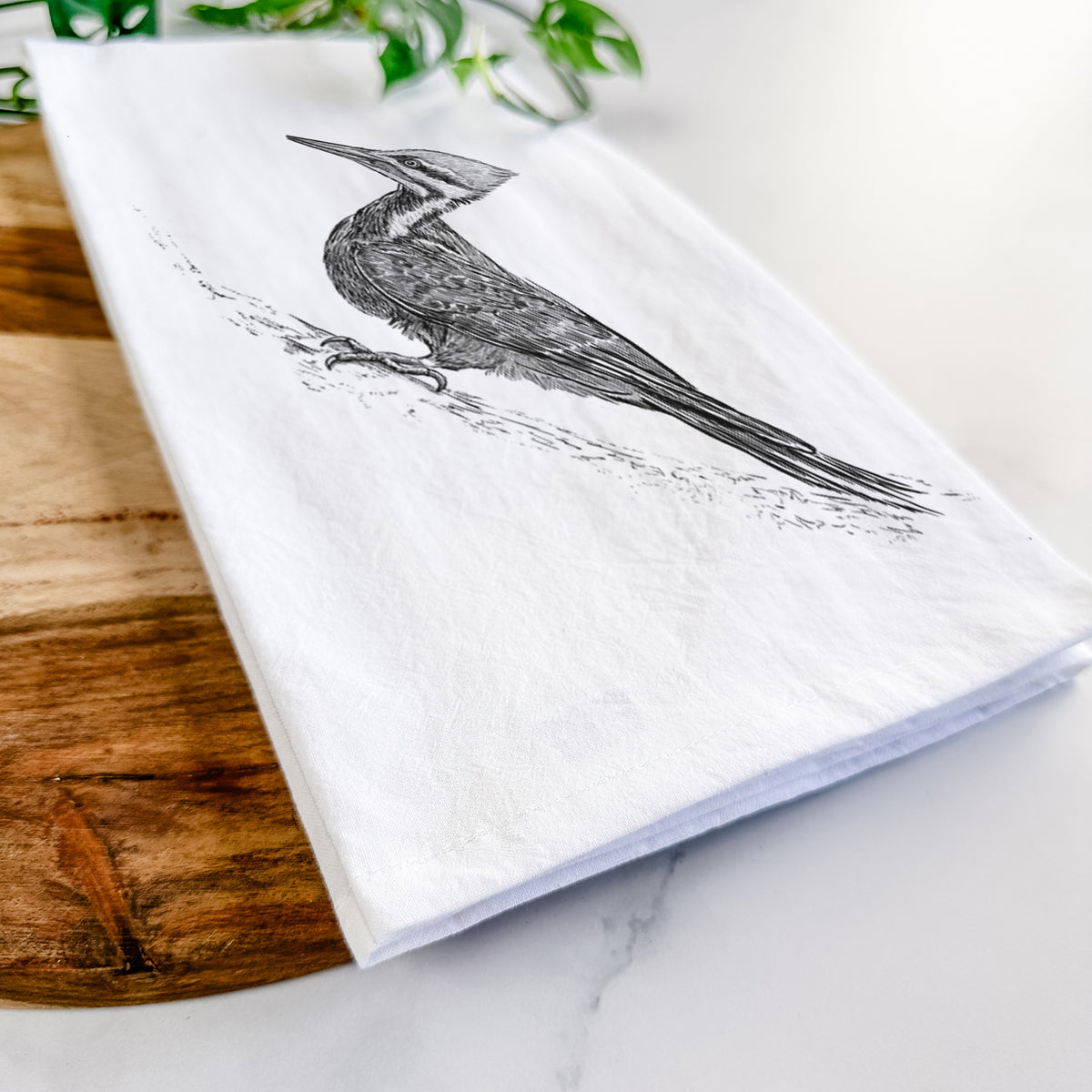 Close-up of a folded white tea towel on a wooden board, showing a black ink illustration of a pileated woodpecker clinging to a tree trunk.