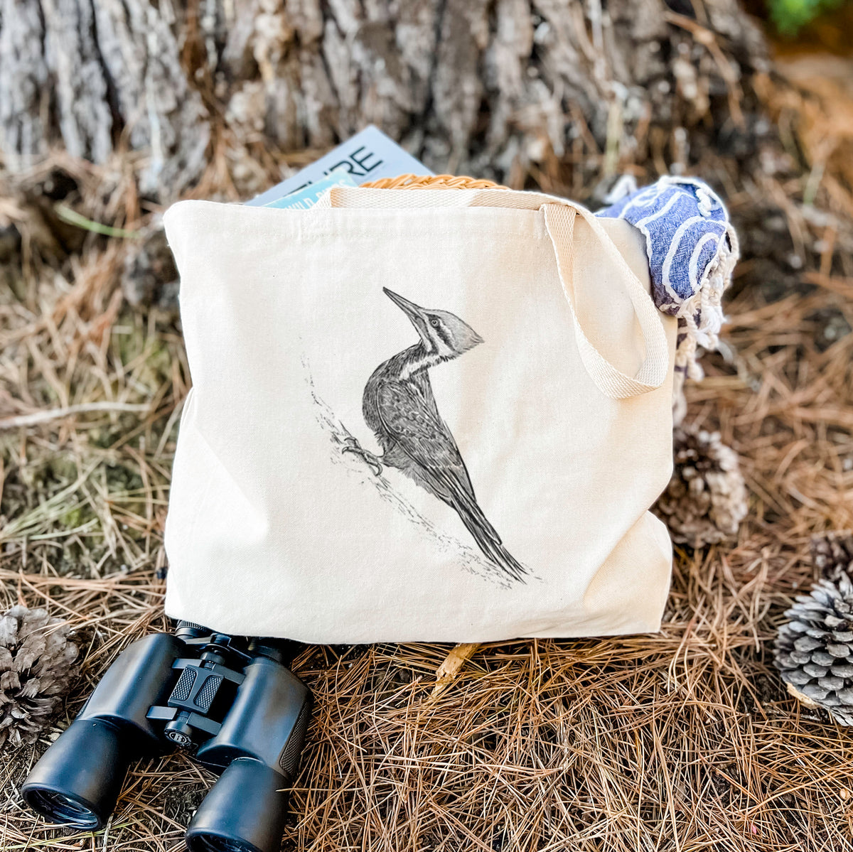 Canvas tote bag printed with a black ink illustration of a pileated woodpecker clinging to a tree trunk, resting on dry grass beside a pair of binoculars.