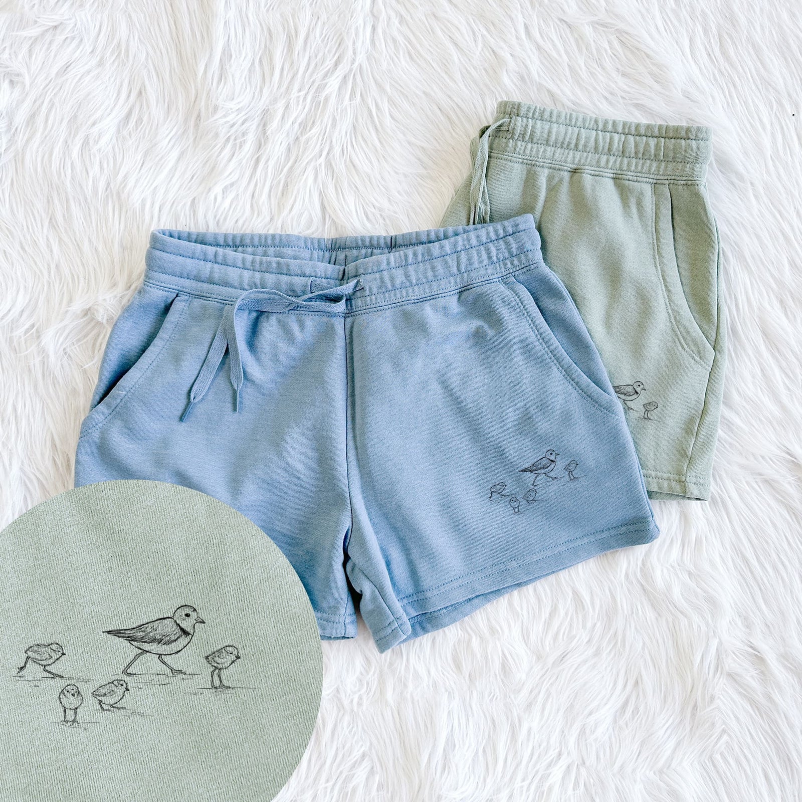 Two pairs of Cali Wave shorts in different colors printed with a black ink sketch of a piping plover with its chicks on the sand, with a close-up of the print.