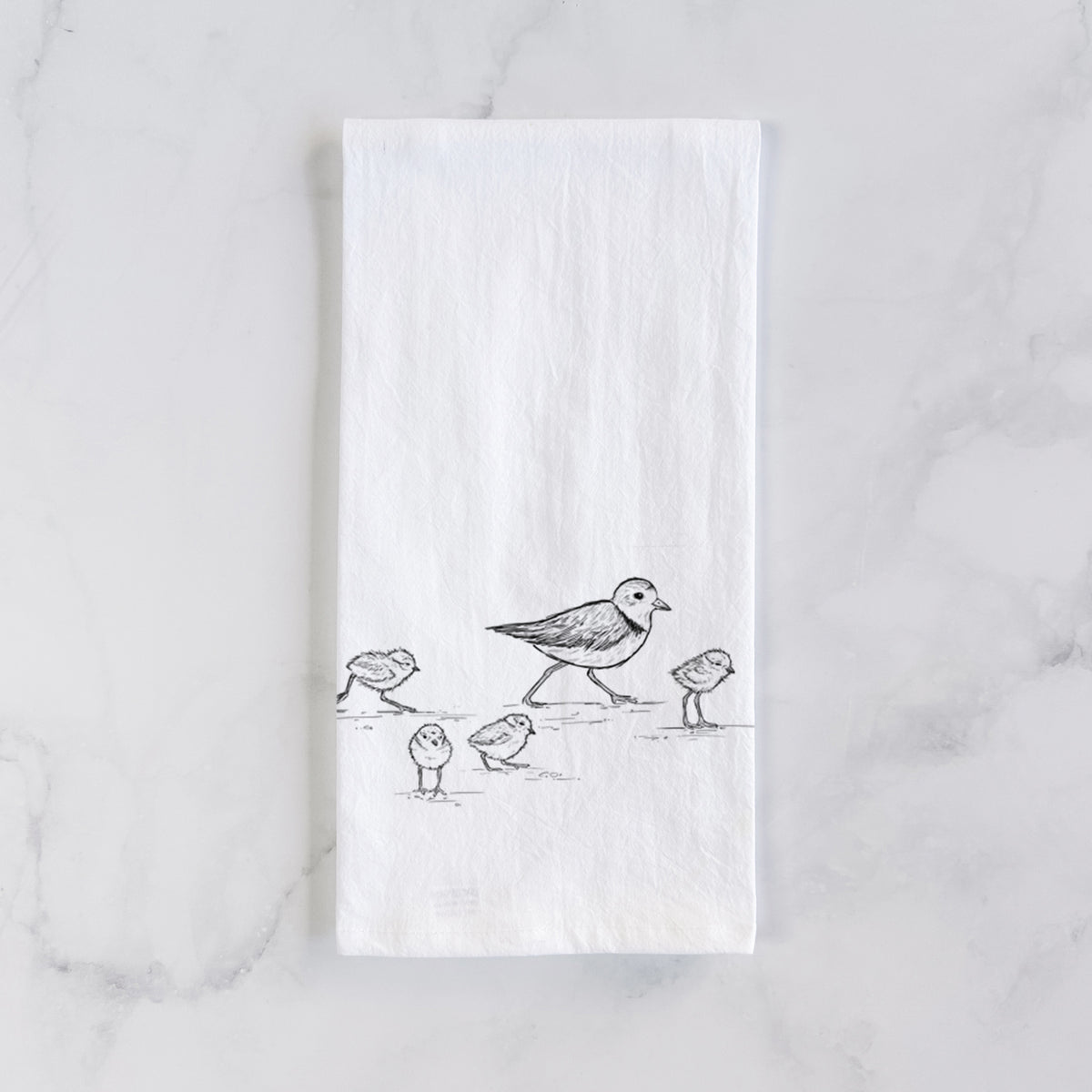 White cotton tea towel laid flat on marble, printed with a black ink sketch of a piping plover with its chicks on the sand.