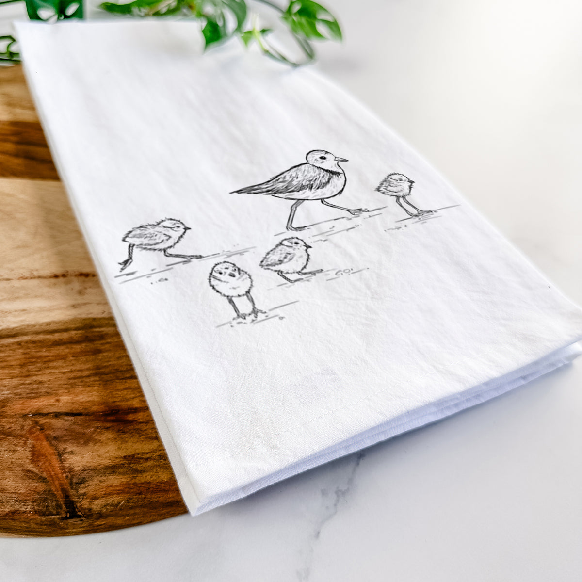 Close-up of a folded white tea towel on a wooden board, showing a black ink sketch of a piping plover with its chicks on the sand.