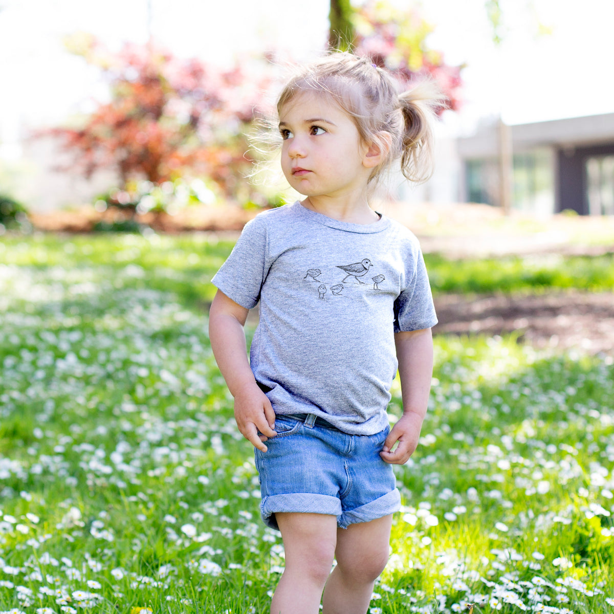 Toddler girl walking outdoors wearing a t-shirt printed with a black ink sketch of a piping plover with its chicks on the sand.
