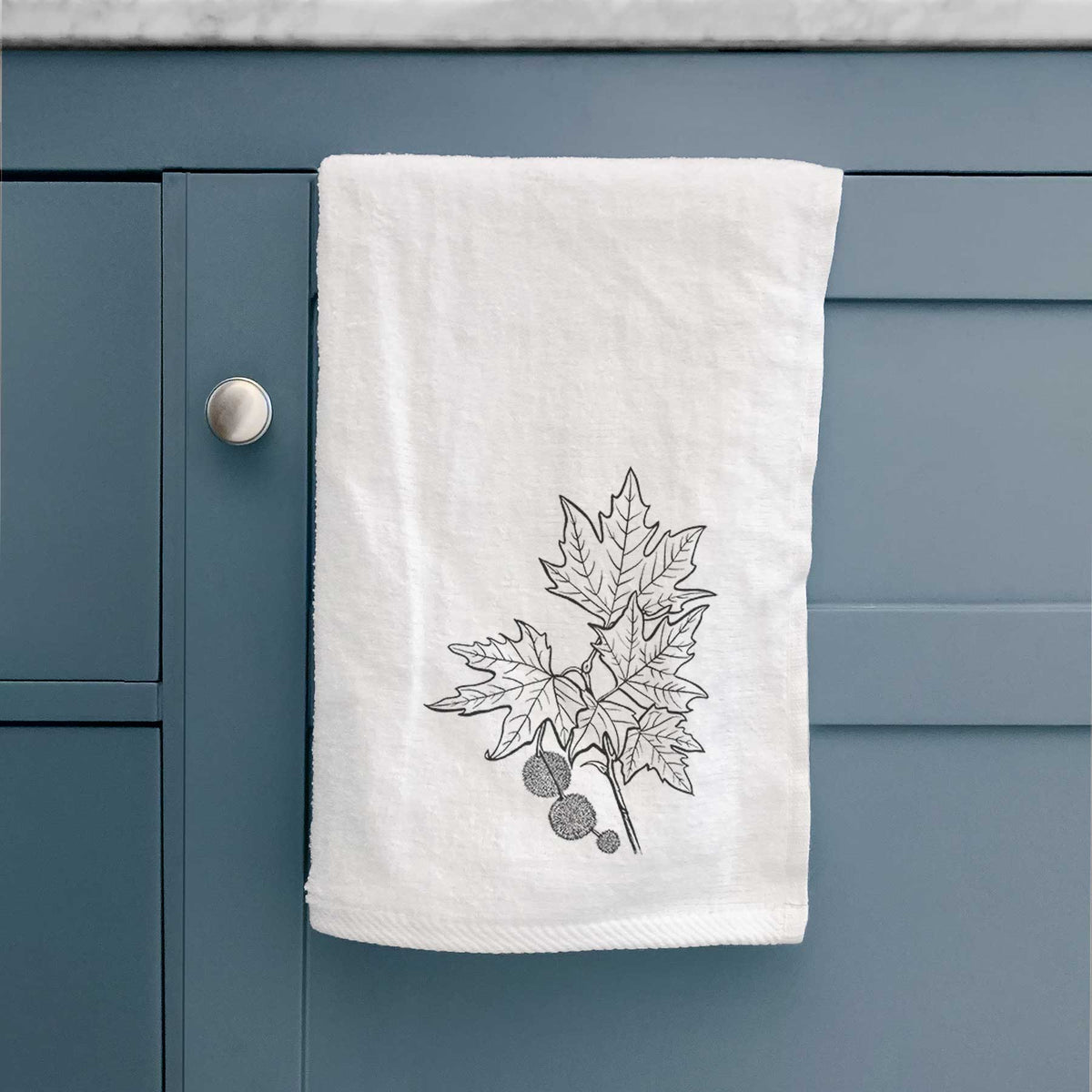 White hand towel printed with a black ink illustration of an oriental plane tree branch with lobed leaves and round seed balls, hanging on a cabinet door.