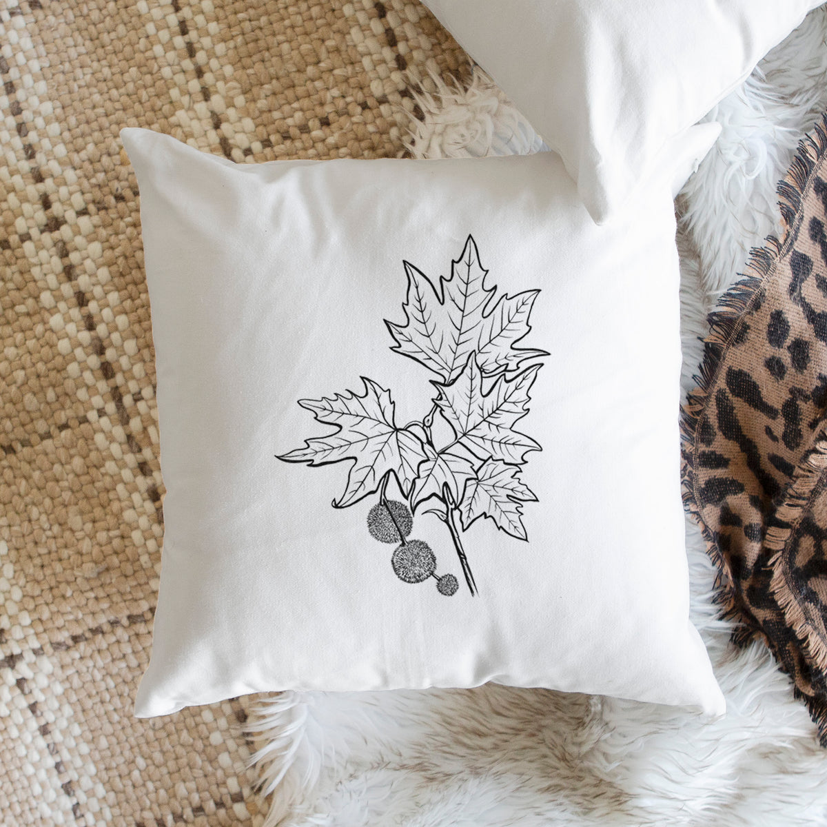 White pillow cover printed with a black ink illustration of an oriental plane tree branch with lobed leaves and round seed balls, on a woven chair.