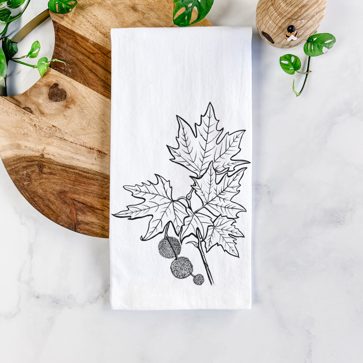 White cotton tea towel printed with a black ink illustration of an oriental plane tree branch with lobed leaves and round seed balls, on a marble counter beside a round wooden cutting board and greenery.