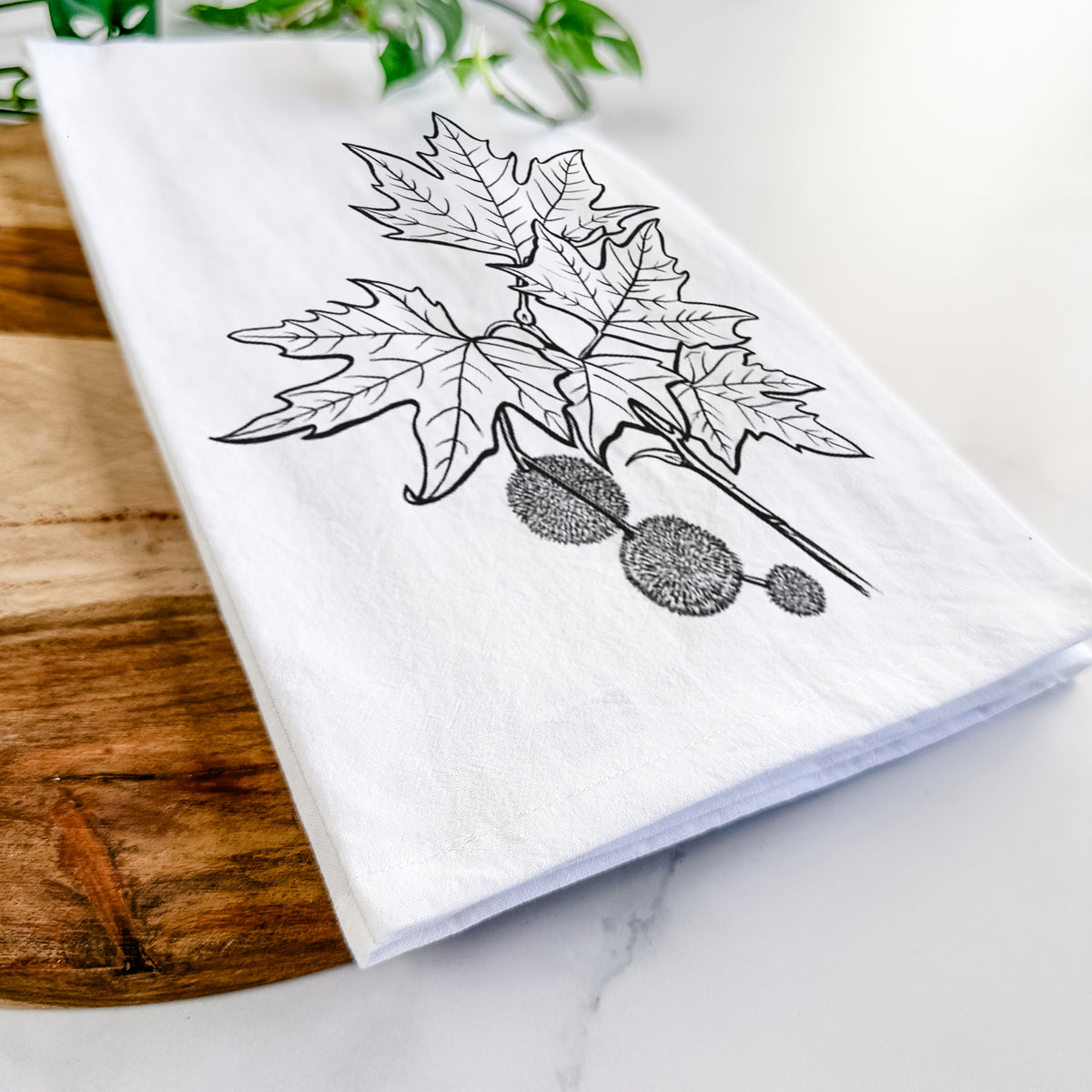 Close-up of a folded white tea towel on a wooden board, showing a black ink illustration of an oriental plane tree branch with lobed leaves and round seed balls.