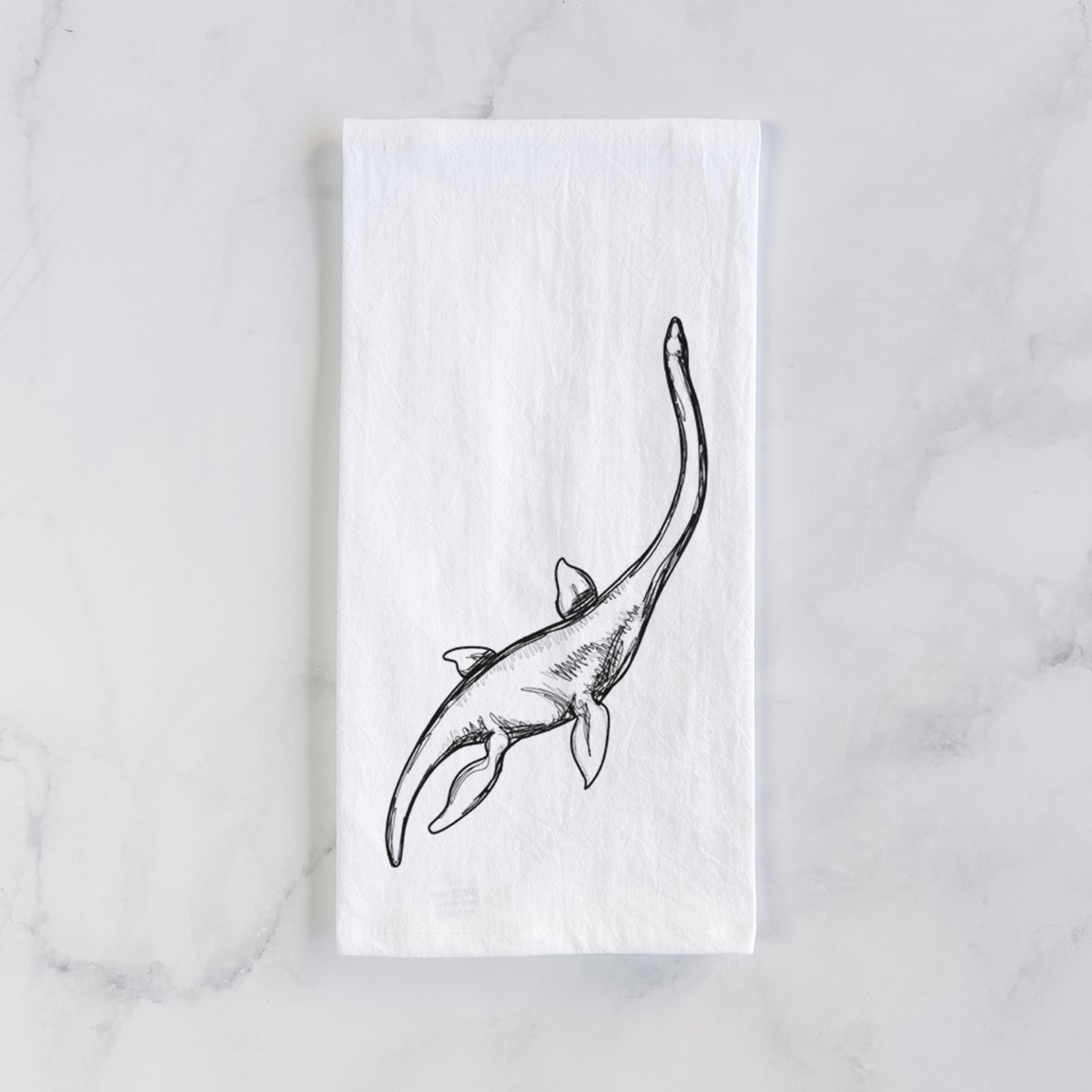 White cotton tea towel printed with a black ink illustration of a long-necked plesiosaur swimming, on a marble counter beside a round wooden cutting board and greenery.