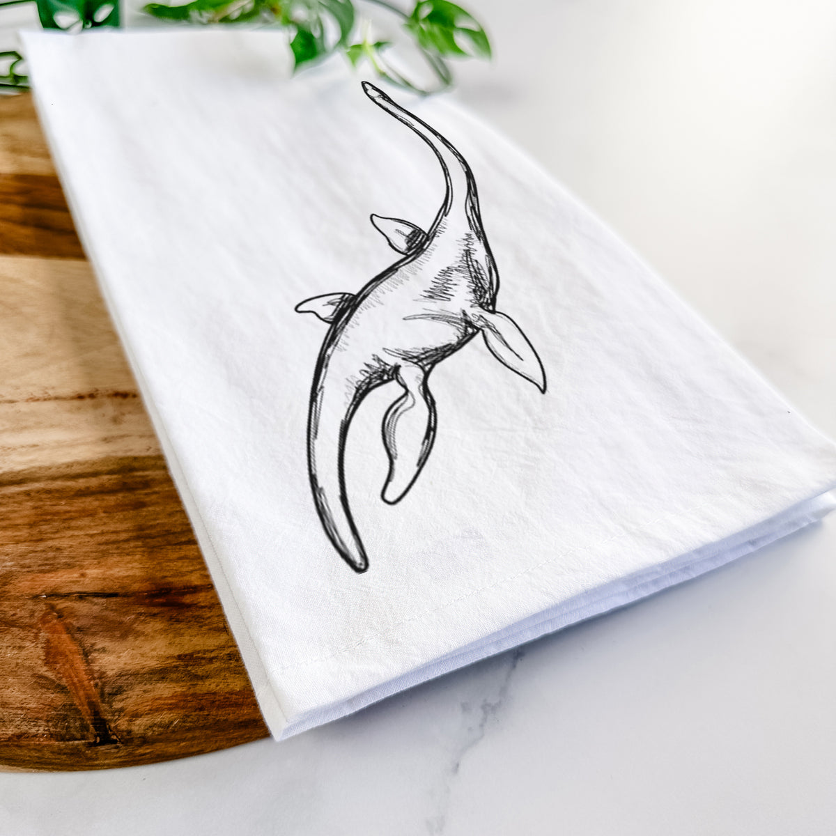 Close-up of a folded white tea towel on a wooden board, showing a black ink illustration of a long-necked plesiosaur swimming.