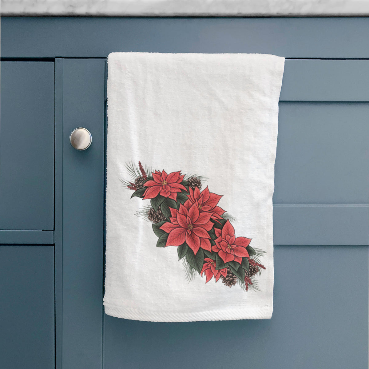White hand towel printed with a full-color illustration of red poinsettia flowers with pine greenery and pine cones, hanging on a cabinet door.