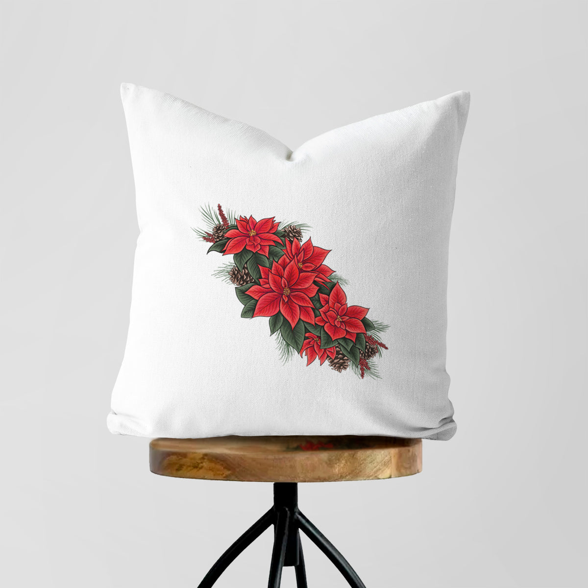 White pillow cover printed with a full-color illustration of red poinsettia flowers with pine greenery and pine cones, on a wooden stool.