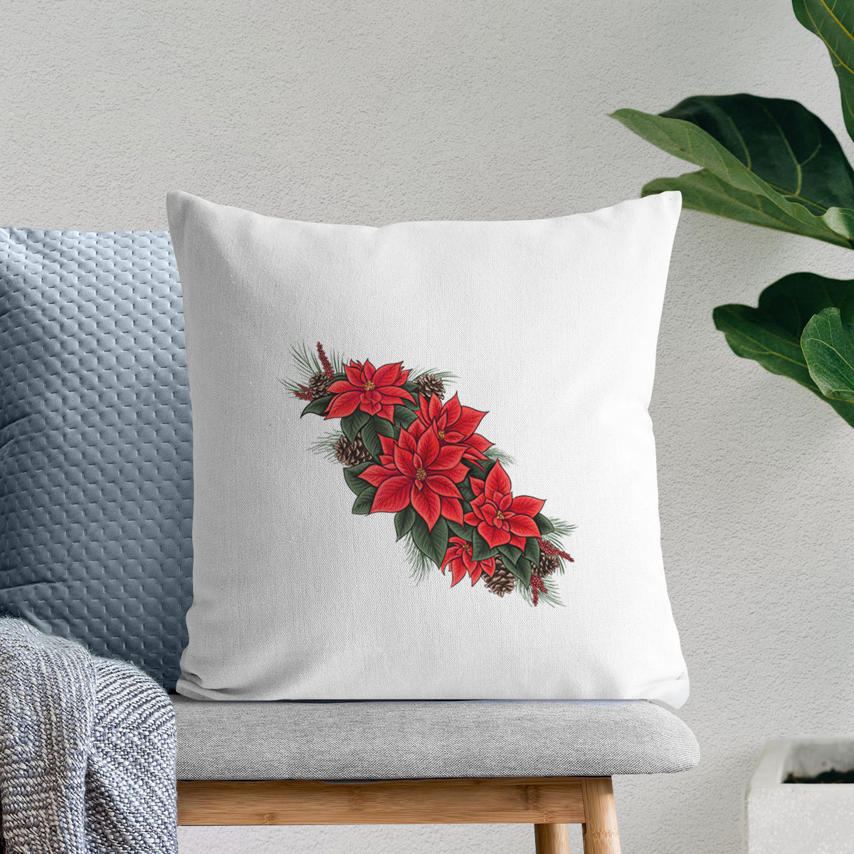 White pillow cover printed with a full-color illustration of red poinsettia flowers with pine greenery and pine cones, on a bench beside plants.