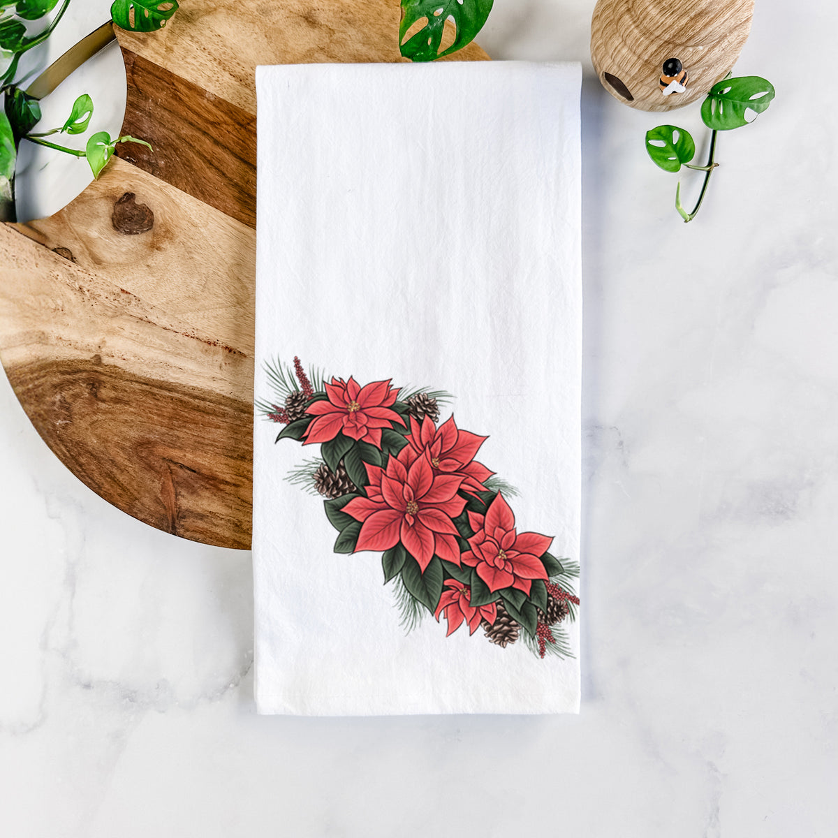 White cotton tea towel printed with a full-color illustration of red poinsettia flowers with pine greenery and pine cones, on a marble counter beside a round wooden cutting board and greenery.
