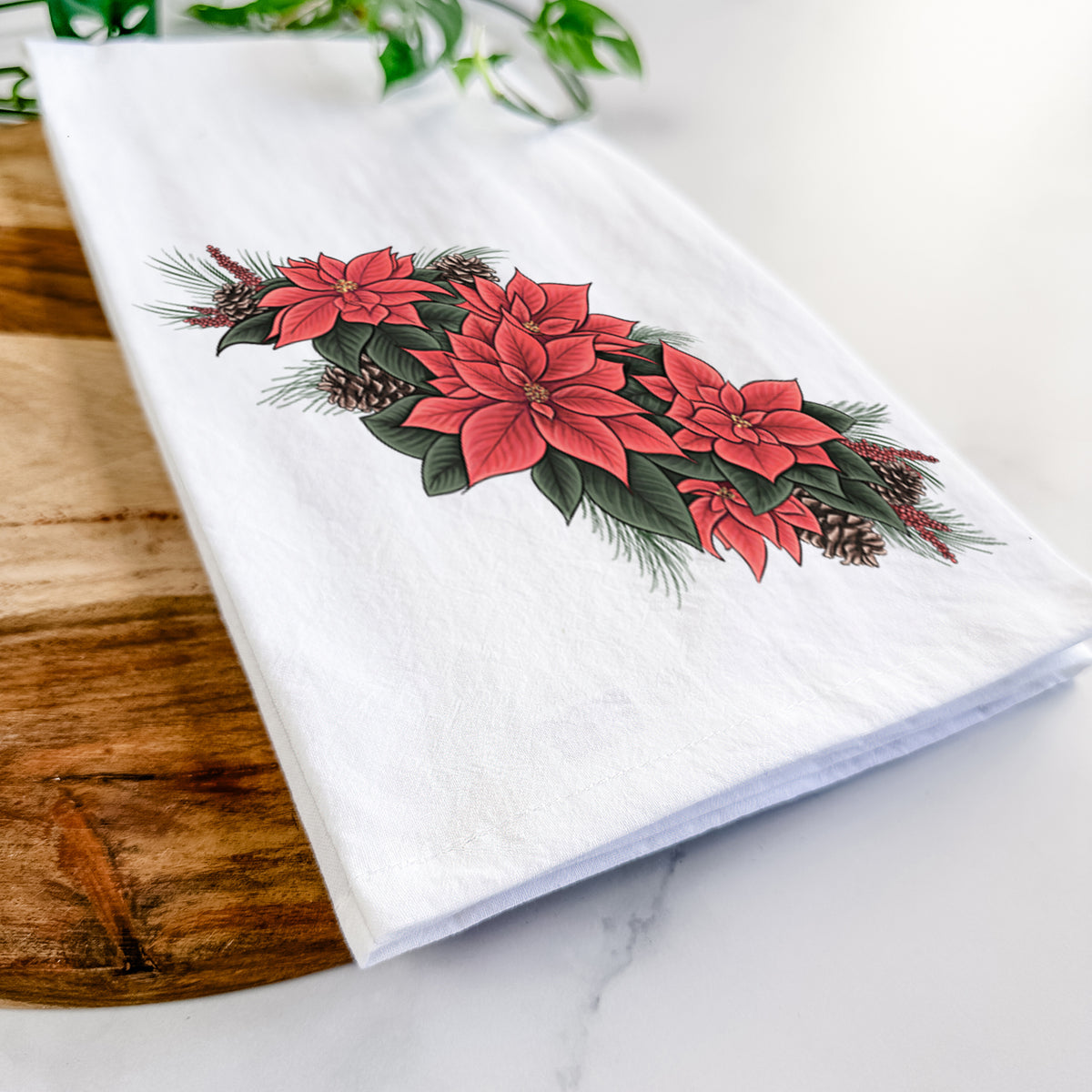 Close-up of a folded white tea towel on a wooden board, showing a full-color illustration of red poinsettia flowers with pine greenery and pine cones.