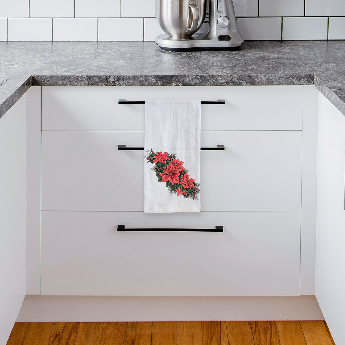 White tea towel printed with a full-color illustration of red poinsettia flowers with pine greenery and pine cones, hanging from a drawer handle in a bright white kitchen.